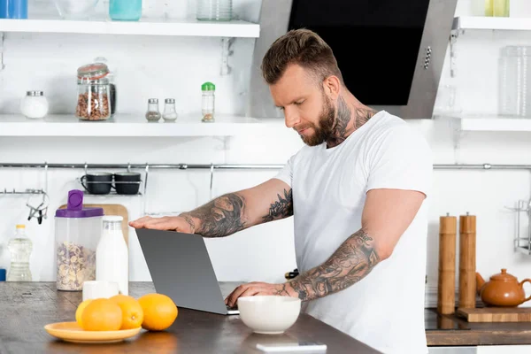 Freelance tatuato in t-shirt bianca con computer portatile vicino a bottiglia di latte, ciotola e arance in cucina — Foto stock