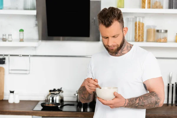 Tätowierter Mann in weißem T-Shirt mixt Frühstück in Schüssel, während er in der Küche steht — Stockfoto