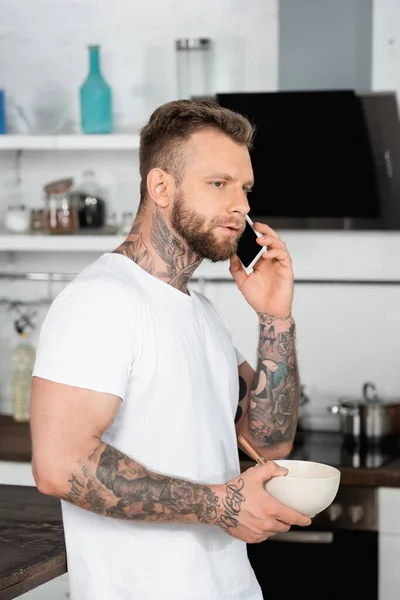 Bearded, tattooed man in white t-shirt holding bowl with breakfast while talking on smartphone in kitchen — Stock Photo