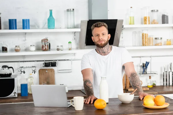 Bearded, tattooed blogger in white t-shirt looking at camera near breakfast and laptop in kitchen — Stock Photo