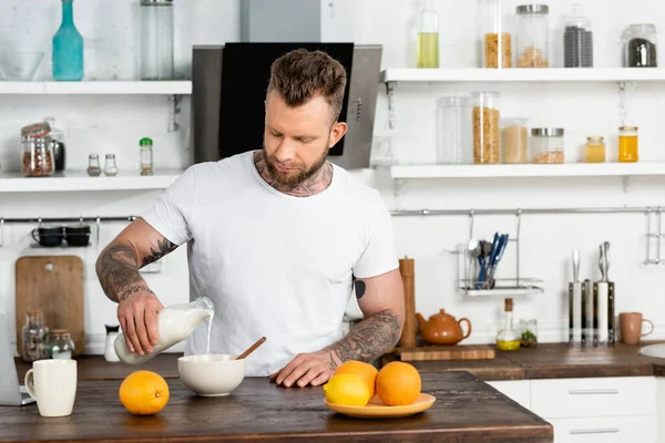 Молодой татуированный мужчина в белой футболке наливает свежее молоко в миску во время завтрака на кухне — стоковое фото