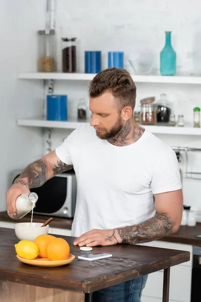 Tattooed man in white t-shirt pouring milk into bowl near oranges and laptop in kitchen — Stock Photo