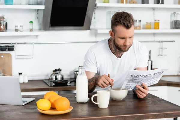 Hombre tatuado en camiseta blanca desayunando en la cocina y leyendo el periódico - foto de stock