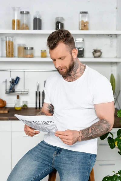 Joven hombre tatuado en camiseta blanca leyendo el periódico en la cocina - foto de stock