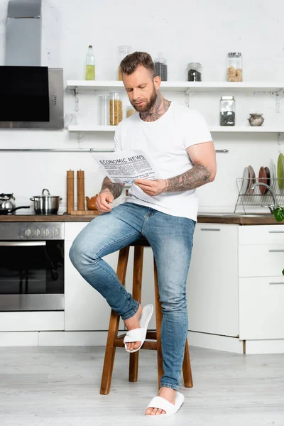 Bearded, tattooed man in white t-shirt and jeans reading newspaper while sitting on stool in kitchen — Stock Photo