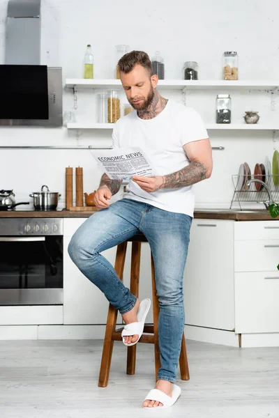 Bärtiger, tätowierter Mann in weißem T-Shirt und Jeans, der Zeitung liest, während er auf einem Hocker in der Küche sitzt — Stockfoto