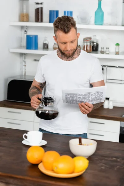 Selective focus of tattooed man in white t-shirt reading newspaper while holding coffee pot in kitchen — Stock Photo