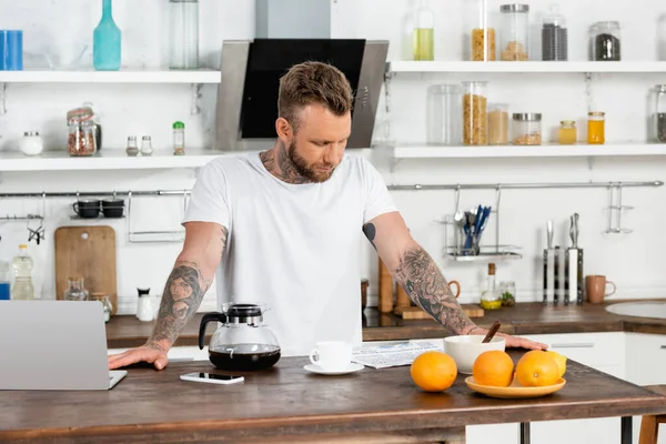 Young man in white t-shirt reading newspaper during breakfast near laptop in kitchen — Stock Photo