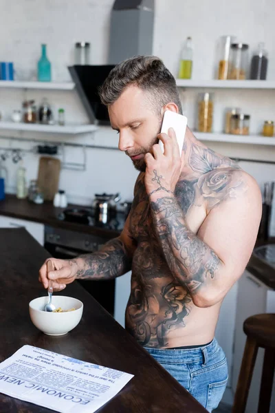 Musculoso, hombre tatuado leyendo periódico y hablando en el teléfono inteligente durante el desayuno en la cocina - foto de stock