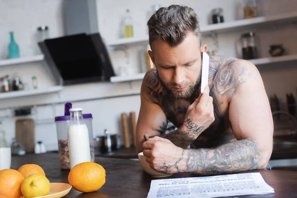 Enfoque selectivo del hombre tatuado sin camisa hablando por teléfono móvil y leyendo el periódico durante el desayuno - foto de stock