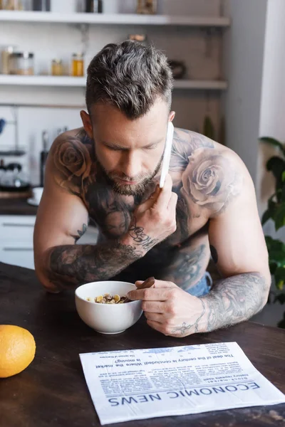 Hemdlos tätowierter Mann spricht während des Frühstücks in Zeitungsnähe auf dem Smartphone — Stockfoto