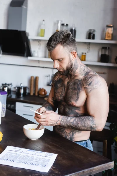 Young tattooed man chatting on smartphone while standing near bowl with breakfast — Stock Photo
