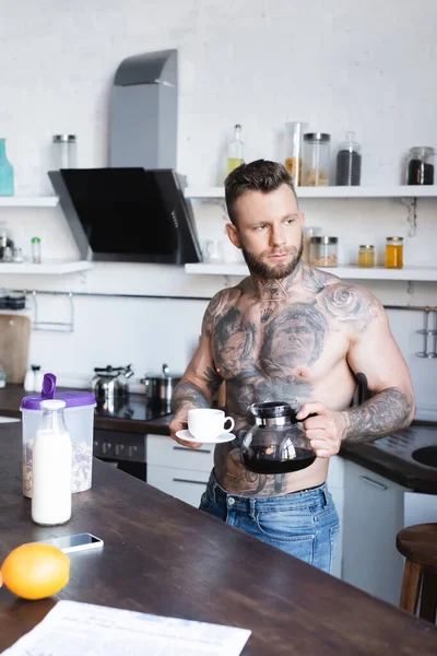 Без рубашки татуированный человек смотрит в сторону, стоя на кухне с кофеваркой и чашкой — стоковое фото