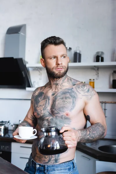 М'язистий, татуйований чоловік тримає каструлю і чашку, стоячи на кухні і дивлячись геть — стокове фото