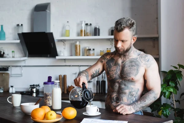 Sin camisa, hombre tatuado vertiendo café en la taza durante el desayuno en la cocina - foto de stock