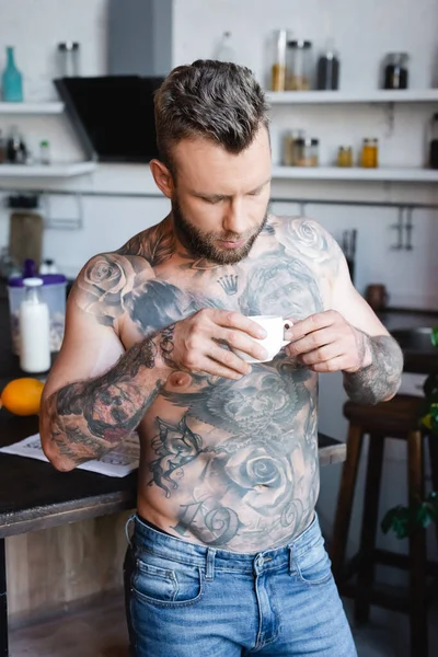 Hombre tatuado sin camisa sosteniendo la taza de café mientras está de pie en la cocina - foto de stock