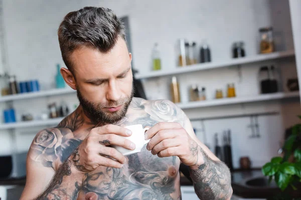 Joven hombre tatuado sosteniendo taza de café en la cocina - foto de stock