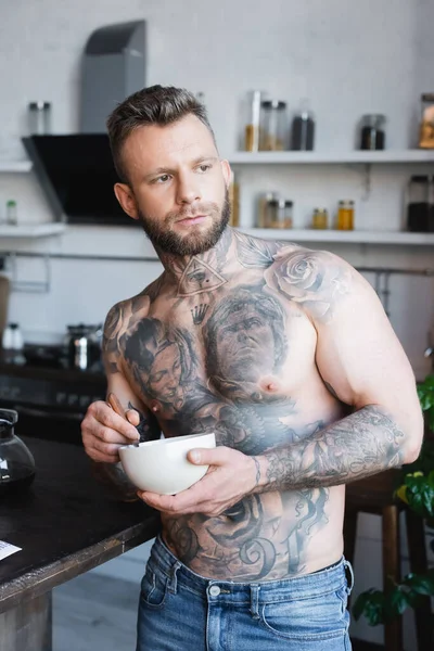 Musclé, tatoué homme regardant loin tout en tenant bol avec petit déjeuner dans la cuisine — Photo de stock