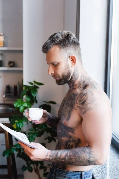 Vue latérale d'un homme tatoué torse nu lisant un journal tout en tenant une tasse de café dans la cuisine — Photo de stock