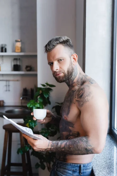 Giovane tatuato che tiene una tazza di caffè e un giornale durante la colazione in cucina — Foto stock