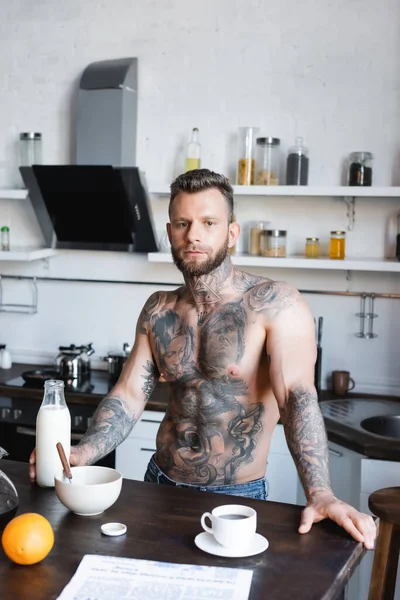 Muscular, tattooed man looking at camera while standing in kitchen during breakfast — Stock Photo