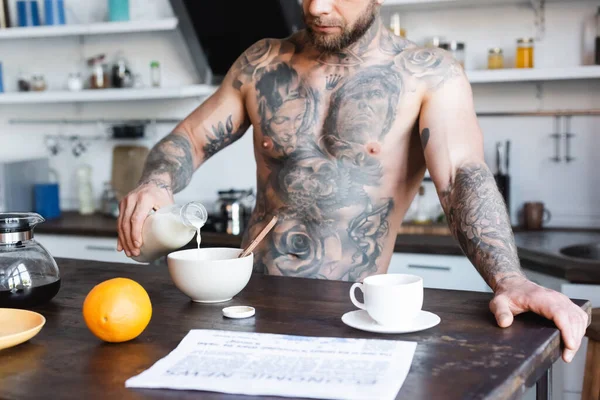 Обрезанный вид татуированного человека, наливающего свежее молоко в миску во время завтрака на кухне возле газеты на столе — стоковое фото