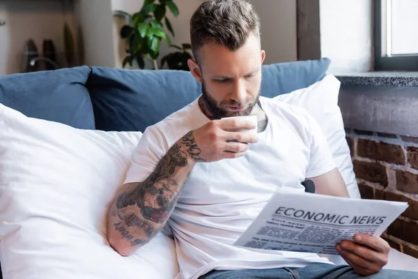 Bärtiger tätowierter Mann trinkt Kaffee und liest Zeitung im Schlafzimmer — Stockfoto