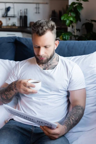 Tattooed man in white t-shirt holding coffee cup while reading newspaper in bedroom — Stock Photo