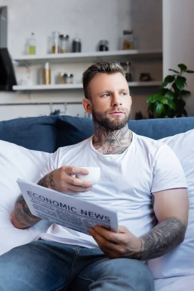 Enfoque selectivo de barbudo hombre tatuado sosteniendo taza de café y periódico mientras está sentado en la cama - foto de stock