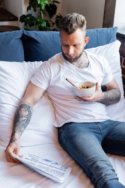 Tattooed man in white t-shirt in jeans reading newspaper while having breakfast in bed — Stock Photo