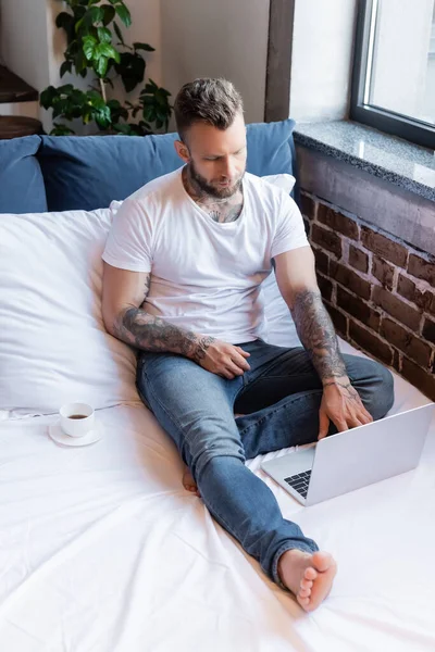 Young tattooed freelancer in jeans and white t-shirt using laptop in bed near cup of coffee — Stock Photo