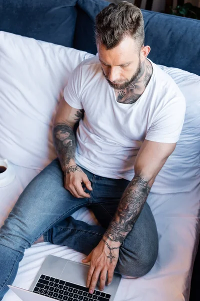 Vista de ángulo alto del freelancer tatuado en camiseta blanca y jeans usando computadora portátil mientras está sentado en la cama - foto de stock