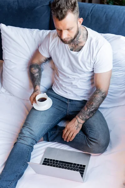 Hochwinkelaufnahme tätowierter Freiberufler in weißem T-Shirt und Jeans mit Laptop und Kaffeetasse im Bett — Stockfoto