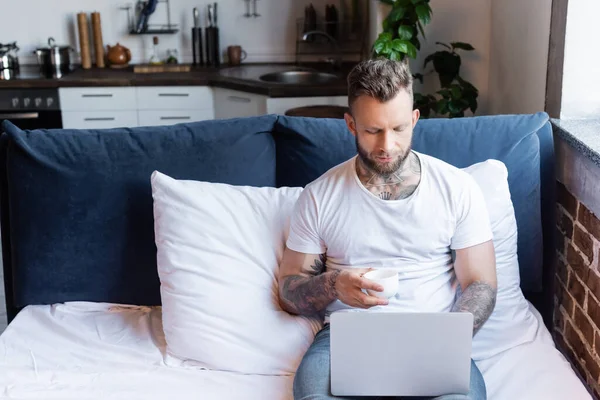 Joven, tatuado freelancer utilizando el ordenador portátil en la cama mientras sostiene la taza de café - foto de stock