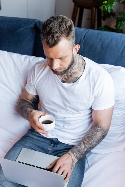 Hochwinkelblick tätowierter Freiberufler, der eine Tasse Kaffee hält, während er Laptop im Schlafzimmer benutzt — Stockfoto