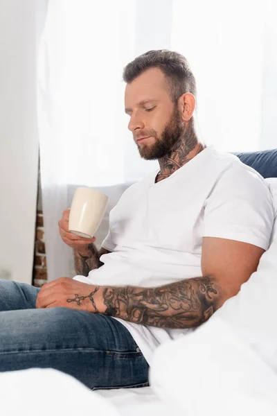 Joven hombre tatuado en camiseta blanca mirando la taza de té mientras está sentado en el dormitorio - foto de stock