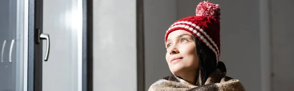 Encabezado del sitio web de la mujer joven en sombrero de punto cálido mirando hacia otro lado mientras está de pie cerca de la ventana en casa - foto de stock