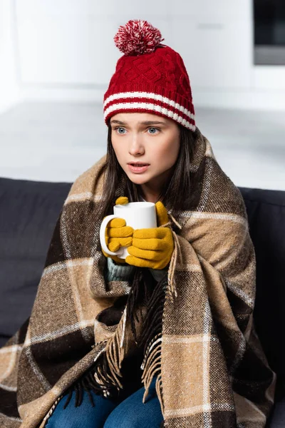 Cold woman, wrapped in plaid blanket, wearing knitted hat and gloves, holding warming drink in kitchen — Stock Photo