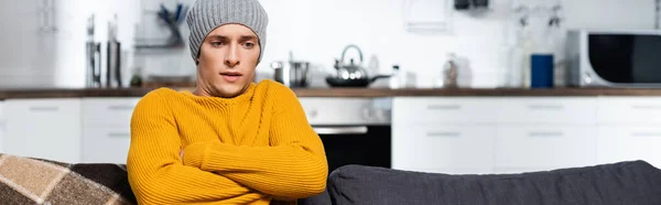 Horizontal crop of freezing man in warm sweater and hat sitting with crossed arms in cold kitchen — Stock Photo