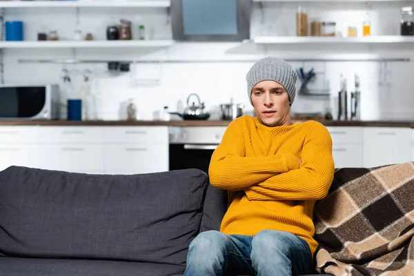 Тремтячий чоловік у в'язаному светрі та капелюсі сидить на дивані з схрещеними руками на холодній кухні — стокове фото