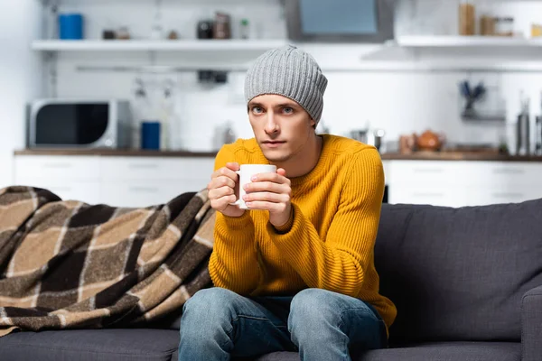 Холодной человек в вязаном свитере и шляпе смотрит в камеру, держа чашку теплого чая на кухне — стоковое фото