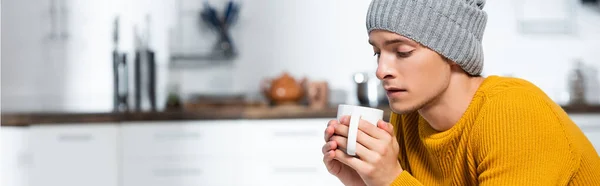 Cabeçalho do site do jovem em chapéu de malha segurando xícara de bebida de aquecimento na cozinha fria — Fotografia de Stock