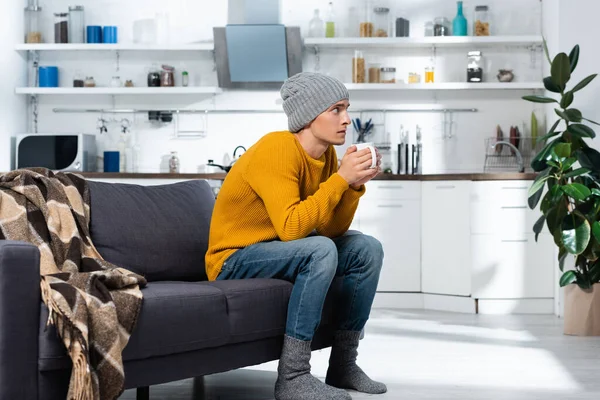 Jeune homme en pull tricoté, chaussettes et chapeau tenant une tasse de thé dans la cuisine — Photo de stock