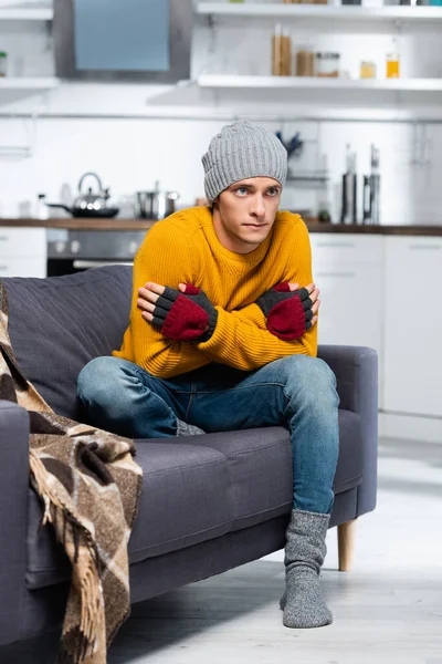 Hombre congelado en sombrero caliente y guantes sin dedos abrazándose mientras está sentado en el sofá en la cocina fría - foto de stock