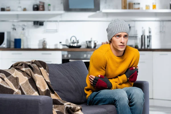 Young man in warm hat and fingerless gloves hugging himself while sitting on sofa in cold kitchen — Stock Photo
