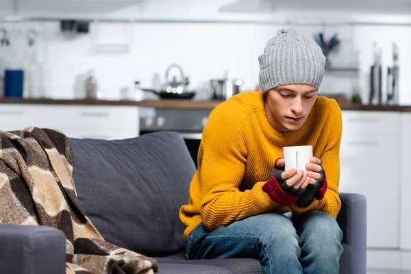 Freezing man in knitted hat and fingerless gloves holding cup of warming beverage while sitting in cold kitchen — Stock Photo