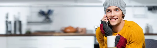 Horizontale Orientierung des jungen Mannes mit Strickmütze und fingerlosen Handschuhen, der mit dem Smartphone spricht, während er eine Tasse warmes Getränk zu Hause hält — Stockfoto