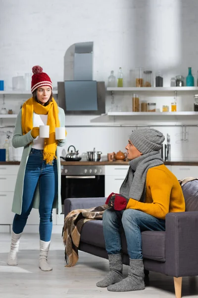 Mulher em roupas quentes carregando copos de bebida quente, enquanto namorado sentado no sofá na cozinha fria — Fotografia de Stock