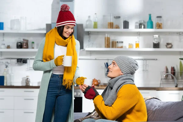 Donna in maglia cappello, sciarpa e guanti che dà tazza di tè caldo al fidanzato seduto sul divano in cucina fredda — Foto stock