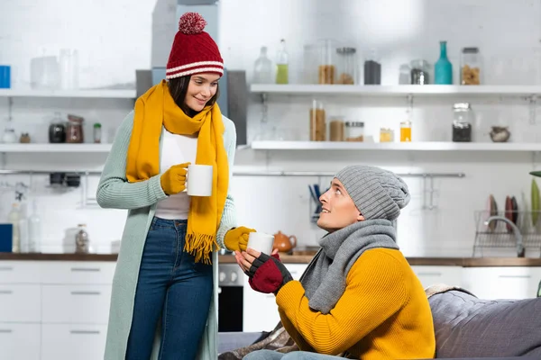 Mujer en sombrero de punto, bufanda y guantes dando taza de té caliente al novio sentado en el sofá en la cocina fría - foto de stock